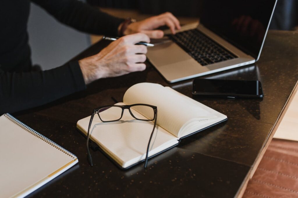 pexels photo 5951722 Professional setting with hands typing on laptop, notebook, and eyeglasses on table.
